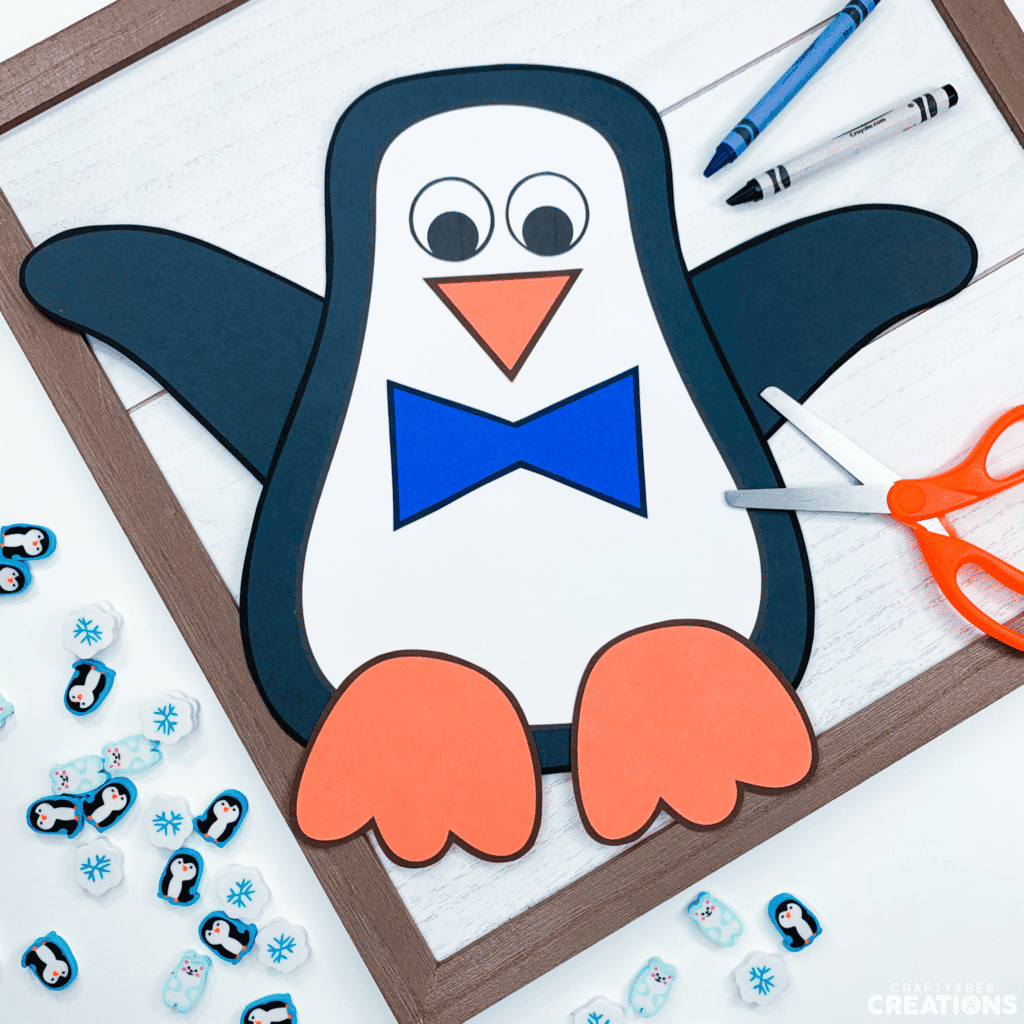 This male penguin craft has a blue bowtie and is lying on a wooden board with orange scissors and a blue and black crayon. The penguins arms are up in the air. There are also mini penguin, snowflake and polar bear erasers on display.