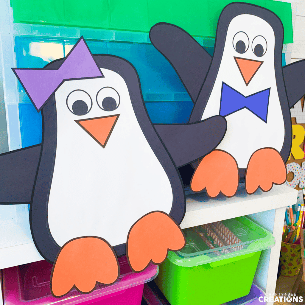 The penguin crafts are hung up in the classroom on some blue and green bins. The penguins have their arms in the air. One penguin has a blue bowtie and the other has a purple bow on its head.