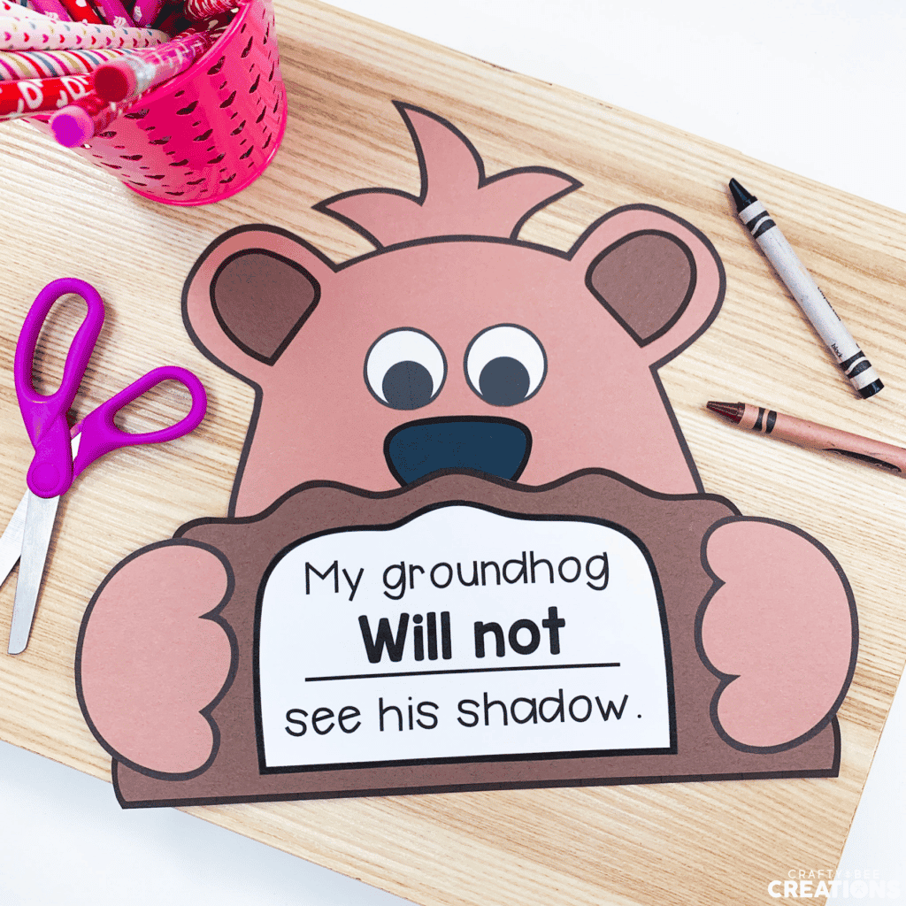 The groundhog craft is displayed on a wooden board with purple scissors and a brown and black crayon. There is a pink pencil cup with Valentine's Day pencils inside.