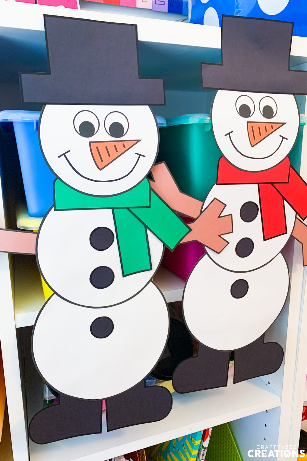 Two snowmen crafts are hung on a shelf where blue and green bins are kept. The snowmen have orange carrot noses, black hats, black buttons and brown stick arms. One is wearing a red scarf and the other has a green scarf. They are also wearing black boots.