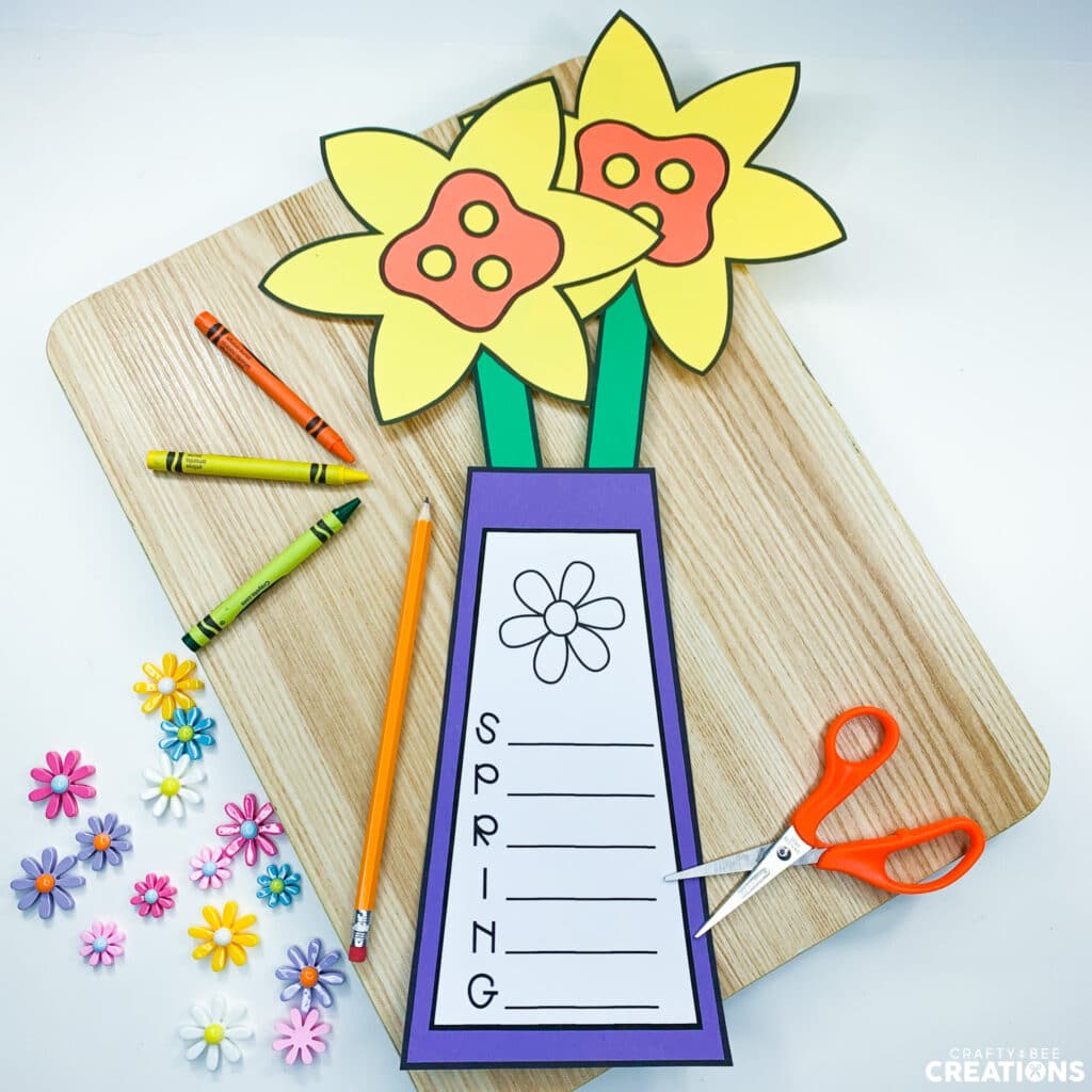 Spring Daffodil Craft is lying on a wooden board with crayons, a pencil, scissors and 3D flowers.