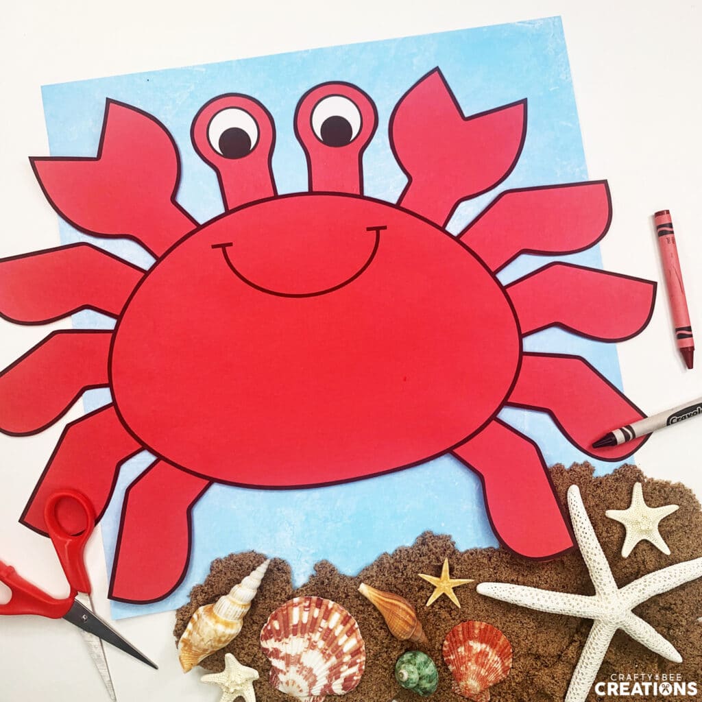 The red crab craft is lying on blue scrapbook paper. There are scissors and crayons nearby. There is also a pile of sand with seashells and a starfish on the page.