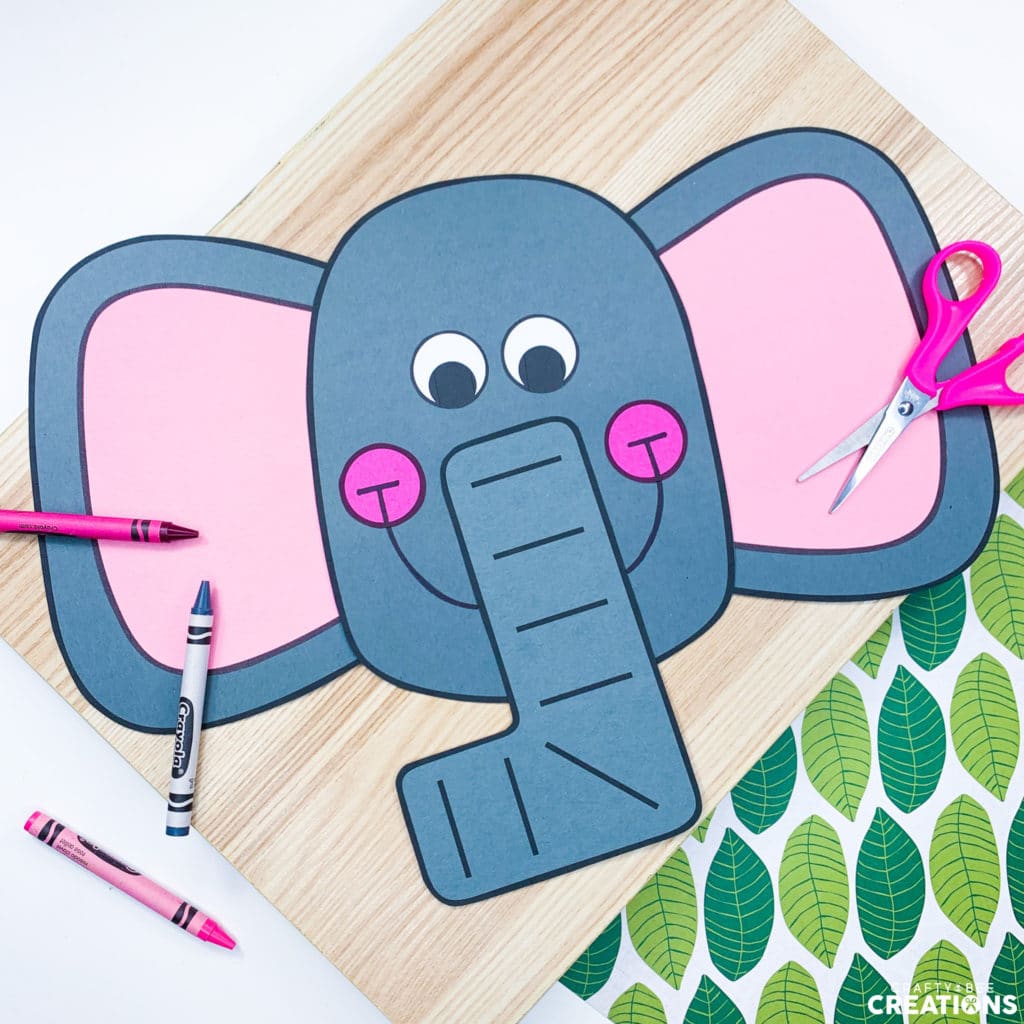 This zoo animal craft is an elephant. The elephant has large gray ears and they are pink on the inside. A pair of pink scissors and some pink and gray crayons lay nearby. The craft is on a wooden board and some green leafy paper.