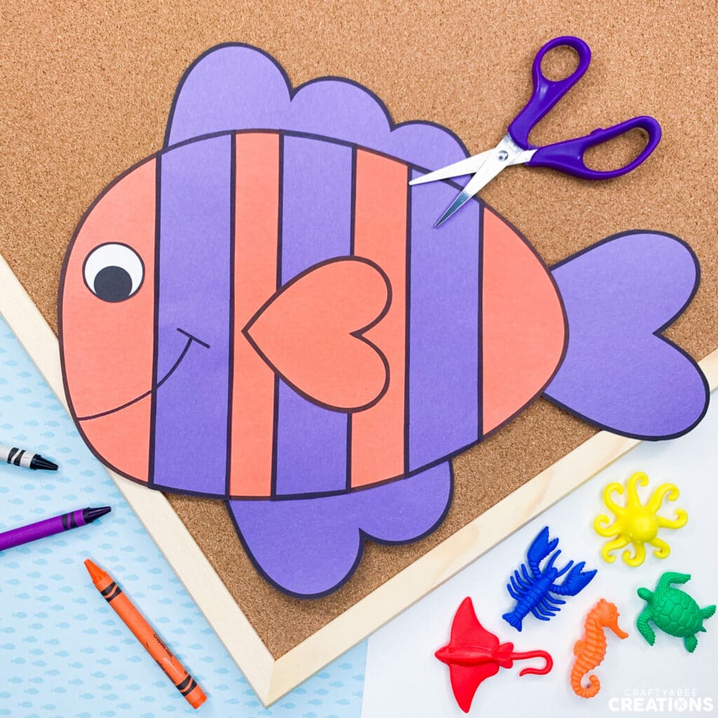 A purple and orange fish craft lies on a corkboard with blue scissors, crayons and some ocean animal figurines.