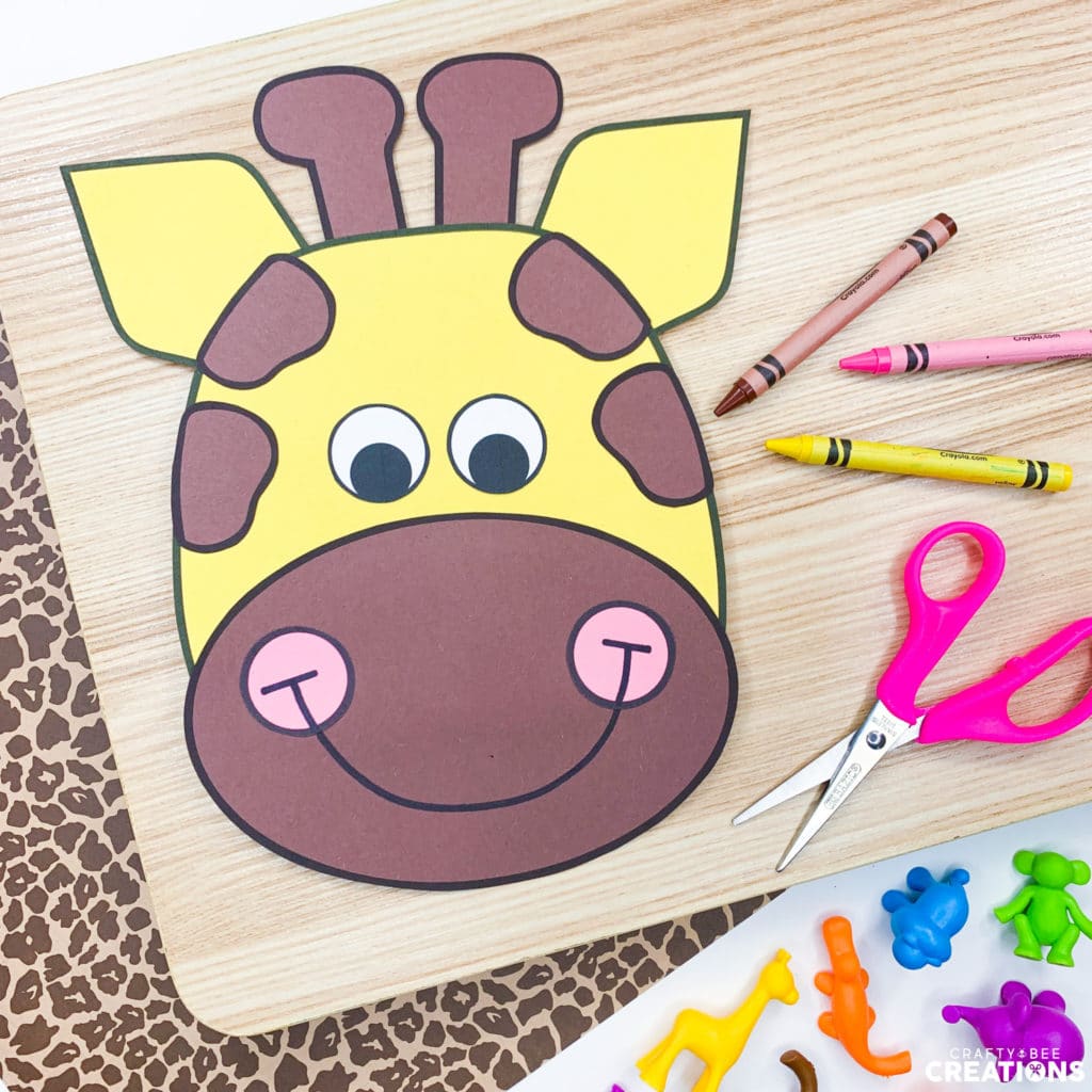 Here is the giraffe zoo animal craft. It is lying on a wooden board and some paper decorated with giraffe spots. There is a brown, yellow and pink crayon and a pink pair of scissors lying nearby. There are also colored animal figurines in the image.