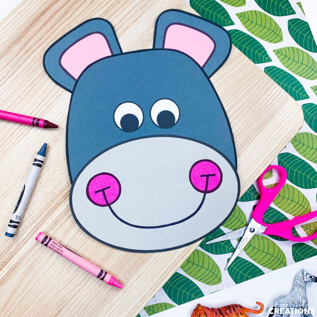 This is a hippo zoo animal craft. The hippo is on a wooden board and some paper decorated with green leaves. There are pink and gray crayons lying near the hippo as well as a pink pair of scissors.