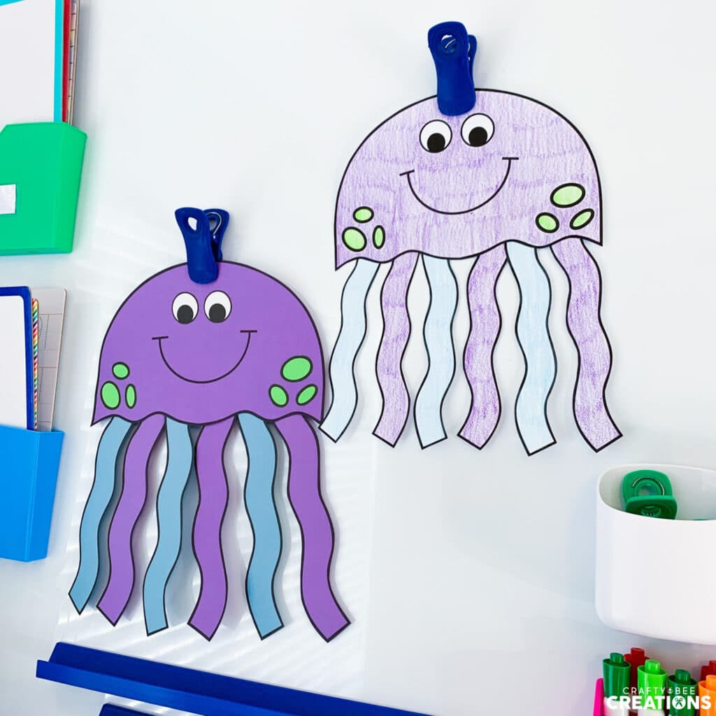 One jellyfish craft is printed on colored cardstock, the other has been colored by a student. They are hanging from magnetic whiteboard clips.