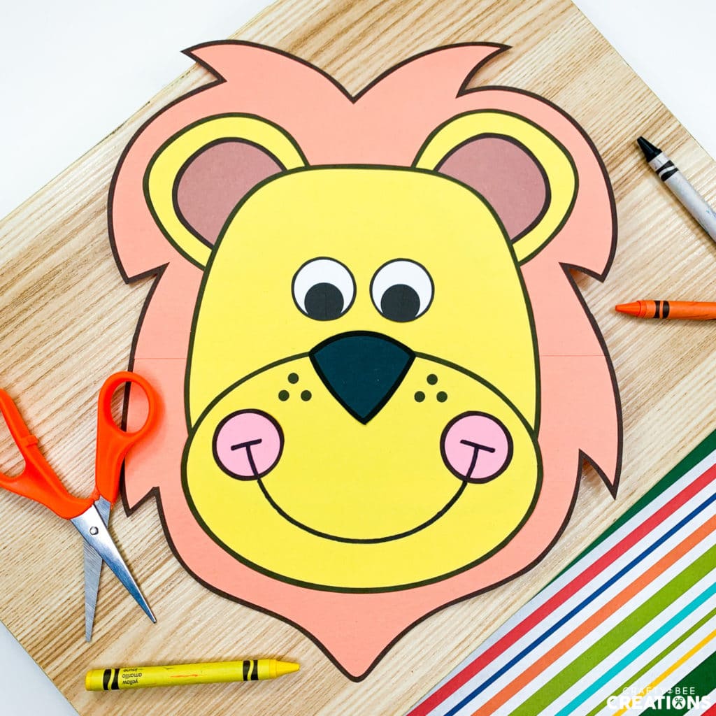 The lion zoo animal craft has been set on a wooden board and a striped piece of paper. The stripes are multicolored. There is a yellow, orange and black crayon on the board, as well as an orange pair of scissors. The lion is yellow with a lion orange mane and rosy pink cheeks.