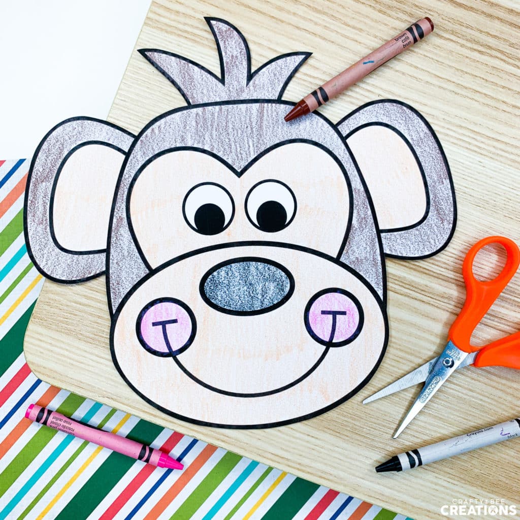This zoo animal craft is the monkey. It has been colored with a brown crayon, which is lying nearby. The craft is on a wooden board and colorful striped paper. There are also orange scissors and a pink and black crayon on the board.