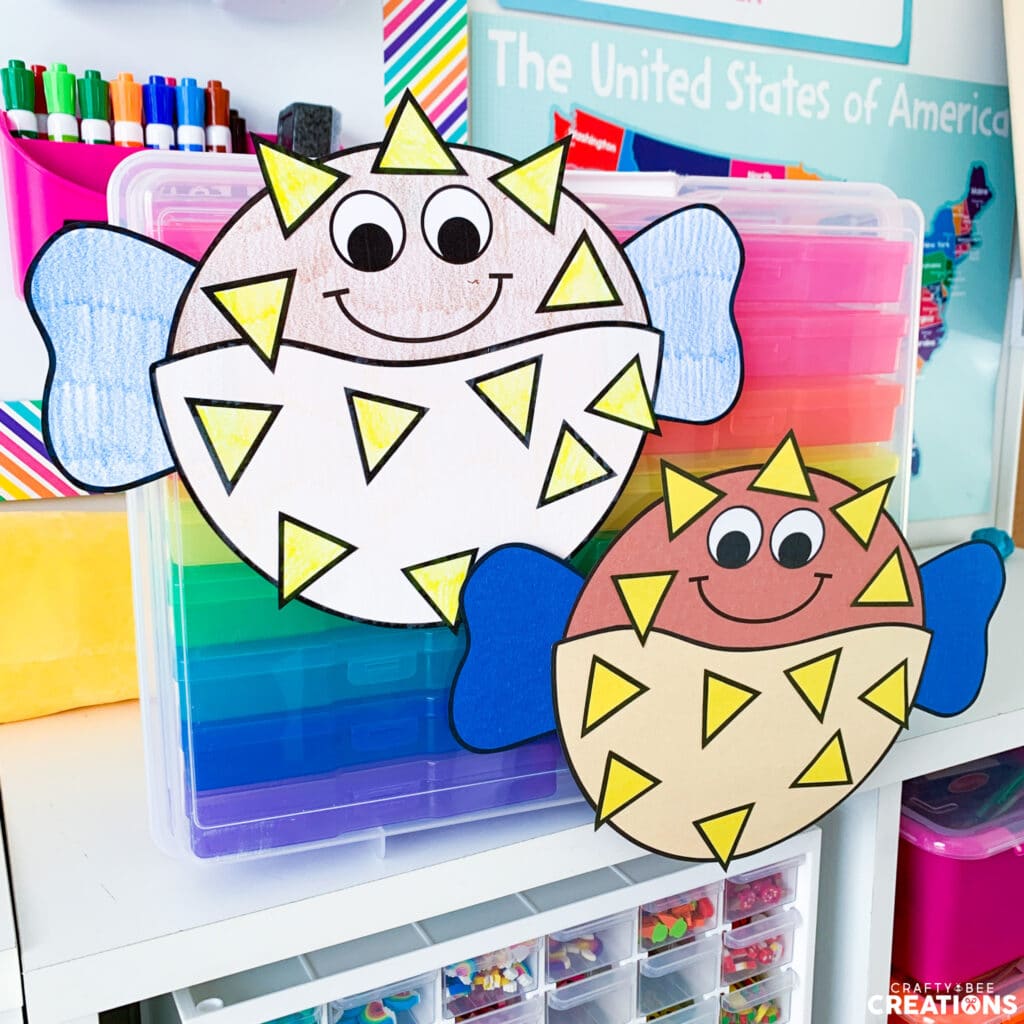 There are two pufferfish crafts. One has been colored by a student and the other has been printed on colored cardstock. They have blue fins and yellow spikes. They are hung on a rainbow case of drawers.