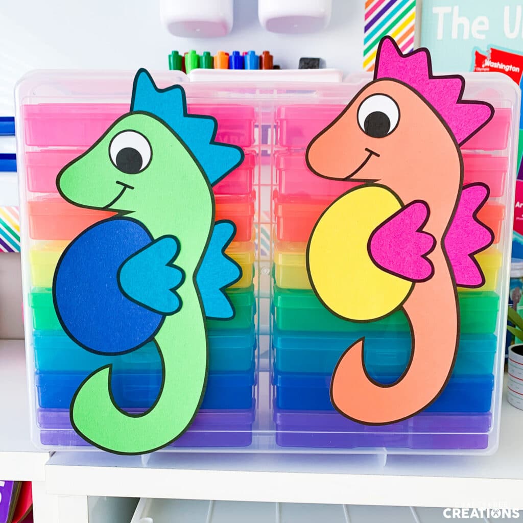 Two seahorses, one green and blue and the other orange, yellow and pink hang on a rainbow set of drawers.