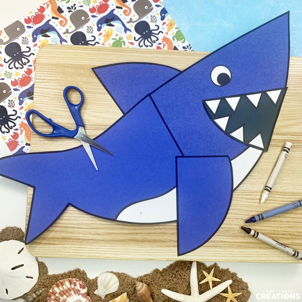 The blue shark craft has many sharp white teeth and is lying on a wooden board. There is sand and seashells as well as ocean animal scrapbook paper nearby.