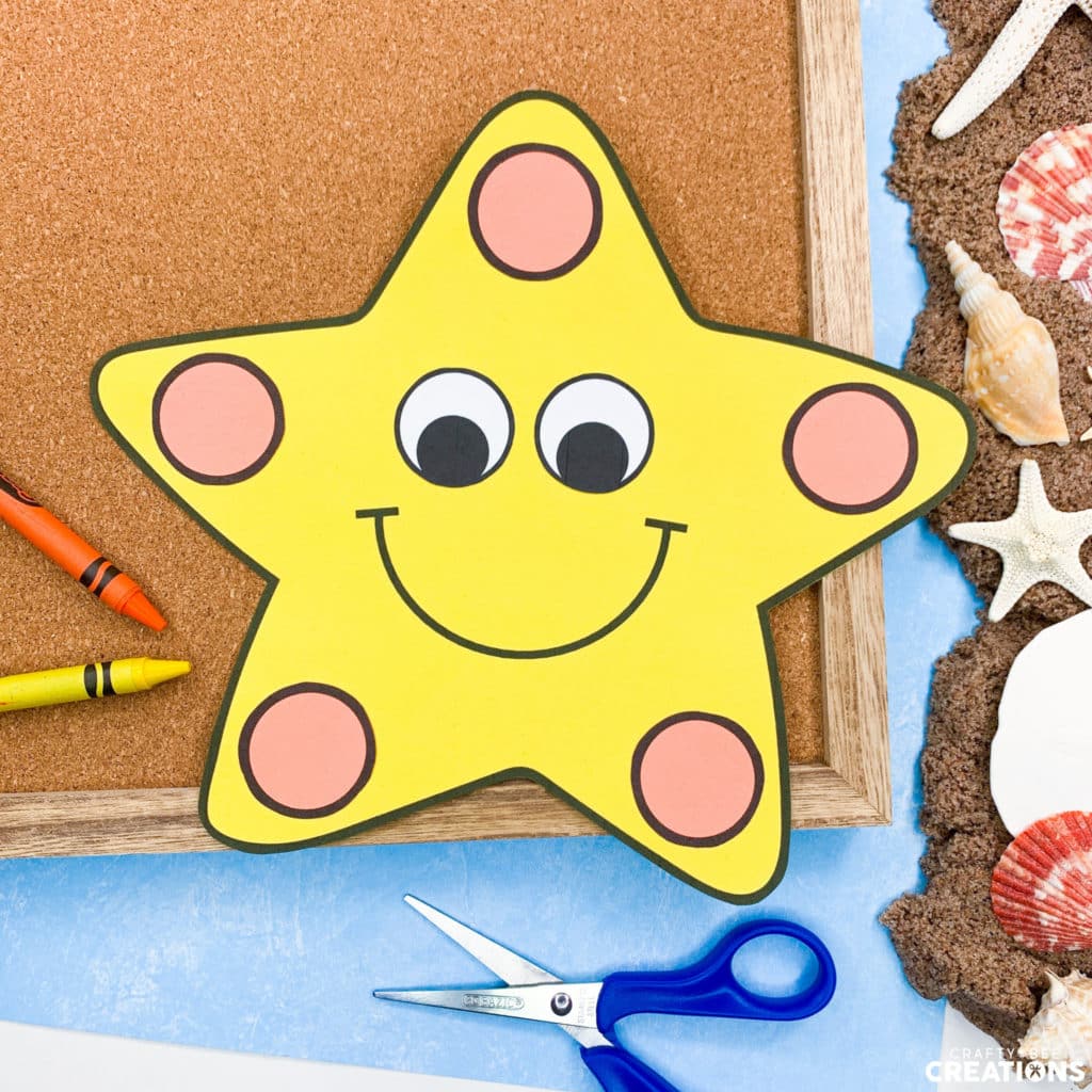 The yellow starfish has orange dots on it's points. It lies on a corkboard with orange and yellow crayons and a blue pair of scissors. There is also a pile of sand and some seashells in the image.