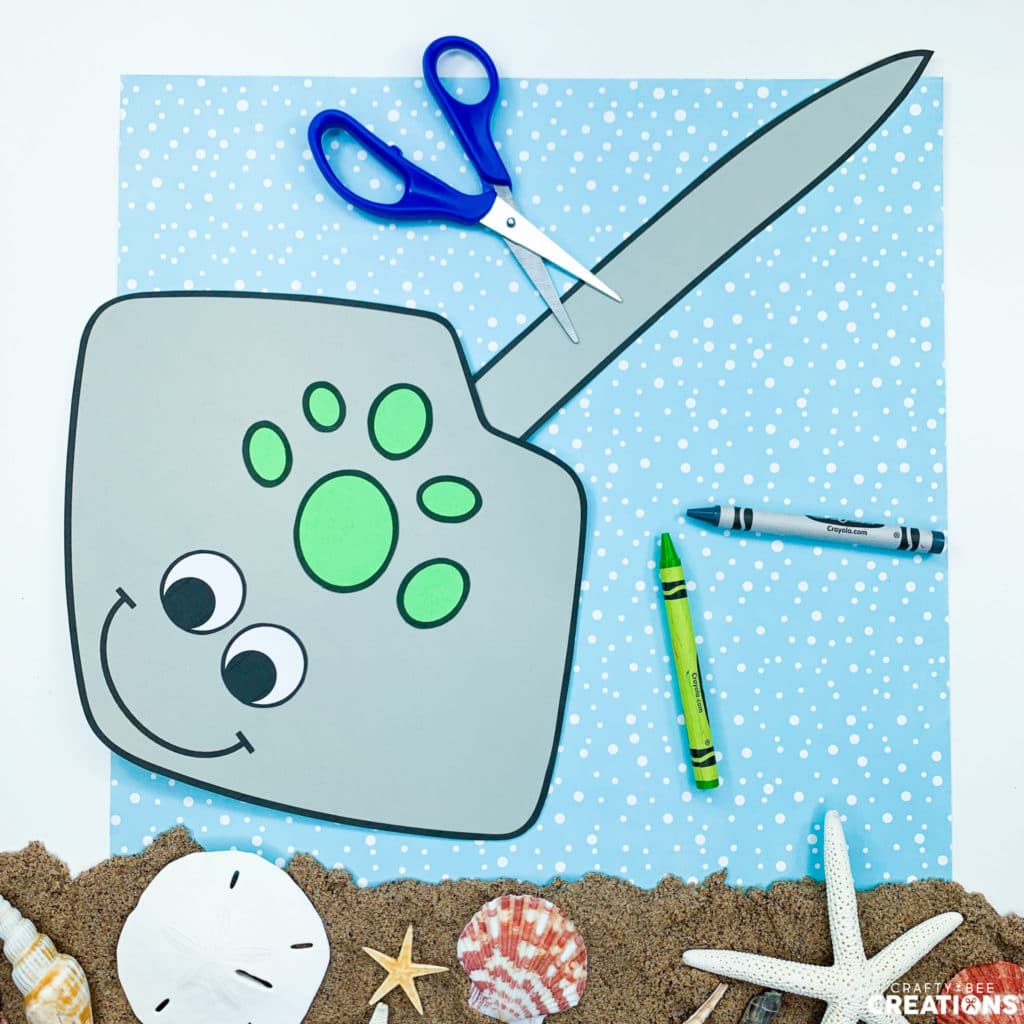 The gray stingray lies on a piece of blue scrapbook paper with blue scissors and crayons. There is a pile of sand and some seashells in the image as well.