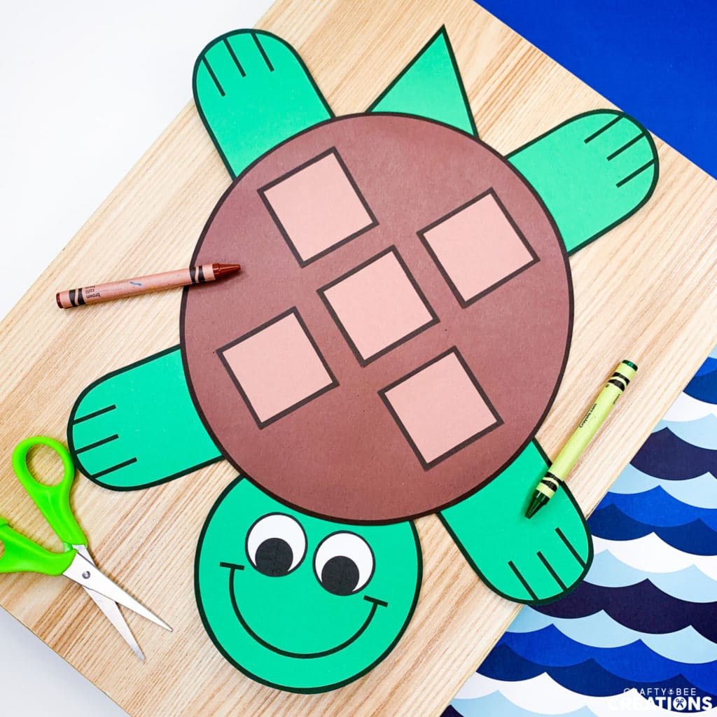 The turtle craft is green and brown and lies on a wooden board. There are brown and green crayons as well as green scissors on the page. The board is on some blue wavy scrapbook paper.