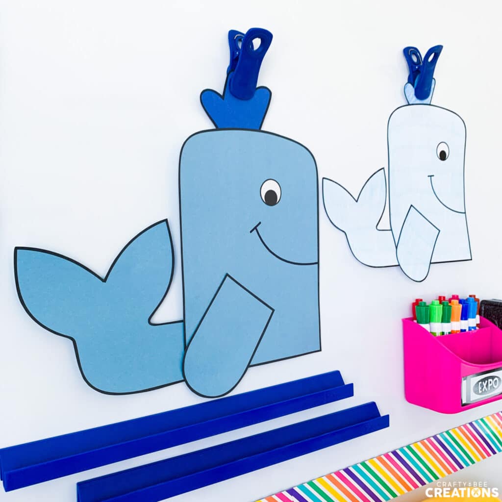 Two whale crafts hang from magnetic clips on a whiteboard. Colorful whiteboard markers are in a cup nearby. One whale is light blue and the other is a darker blue.