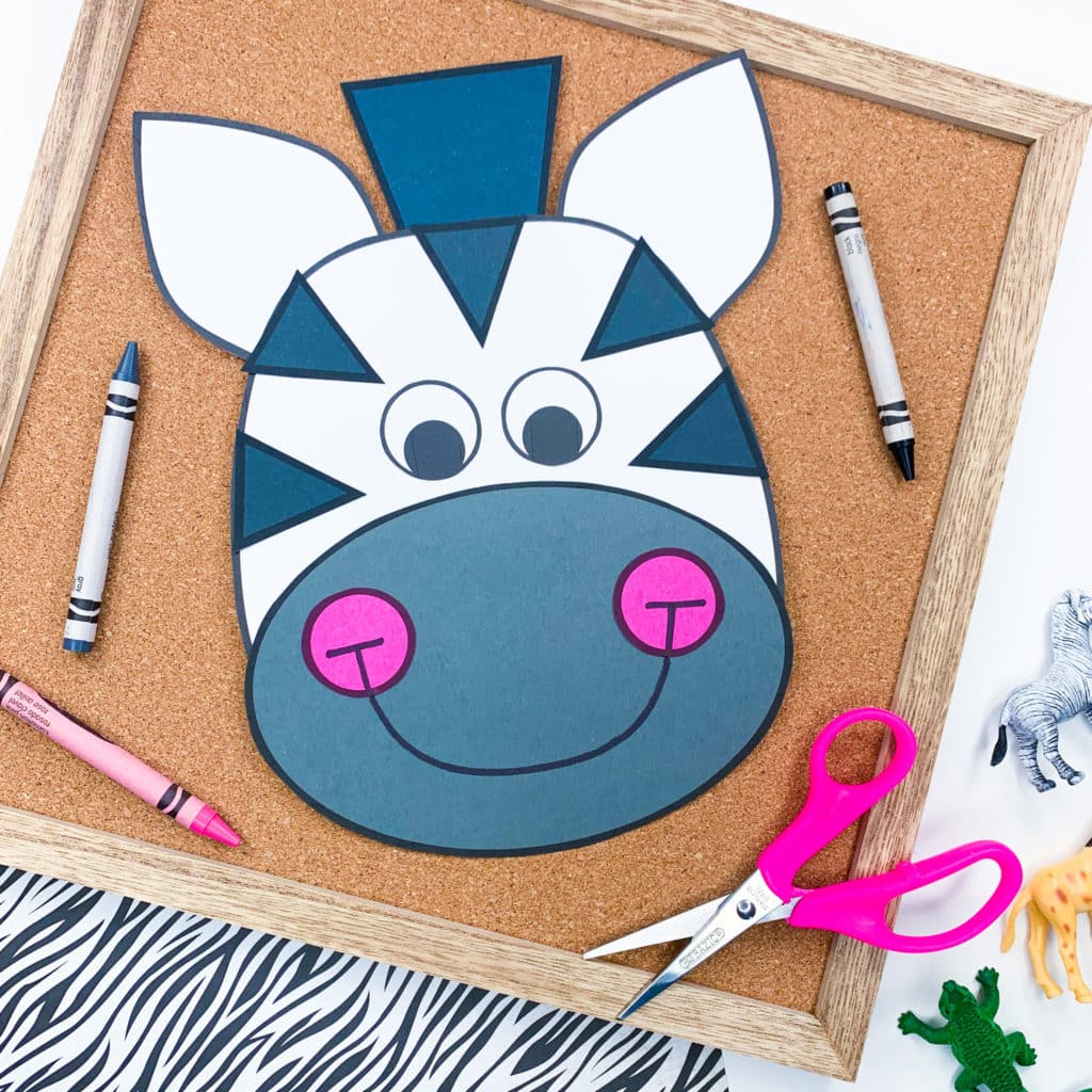 The zebra zoo animal craft has been placed on a brown cork board with a gray, black and pink crayon. There is also a pink pair of scissors. The board is on striped zebra paper. Some small animal figurines are lying nearby.