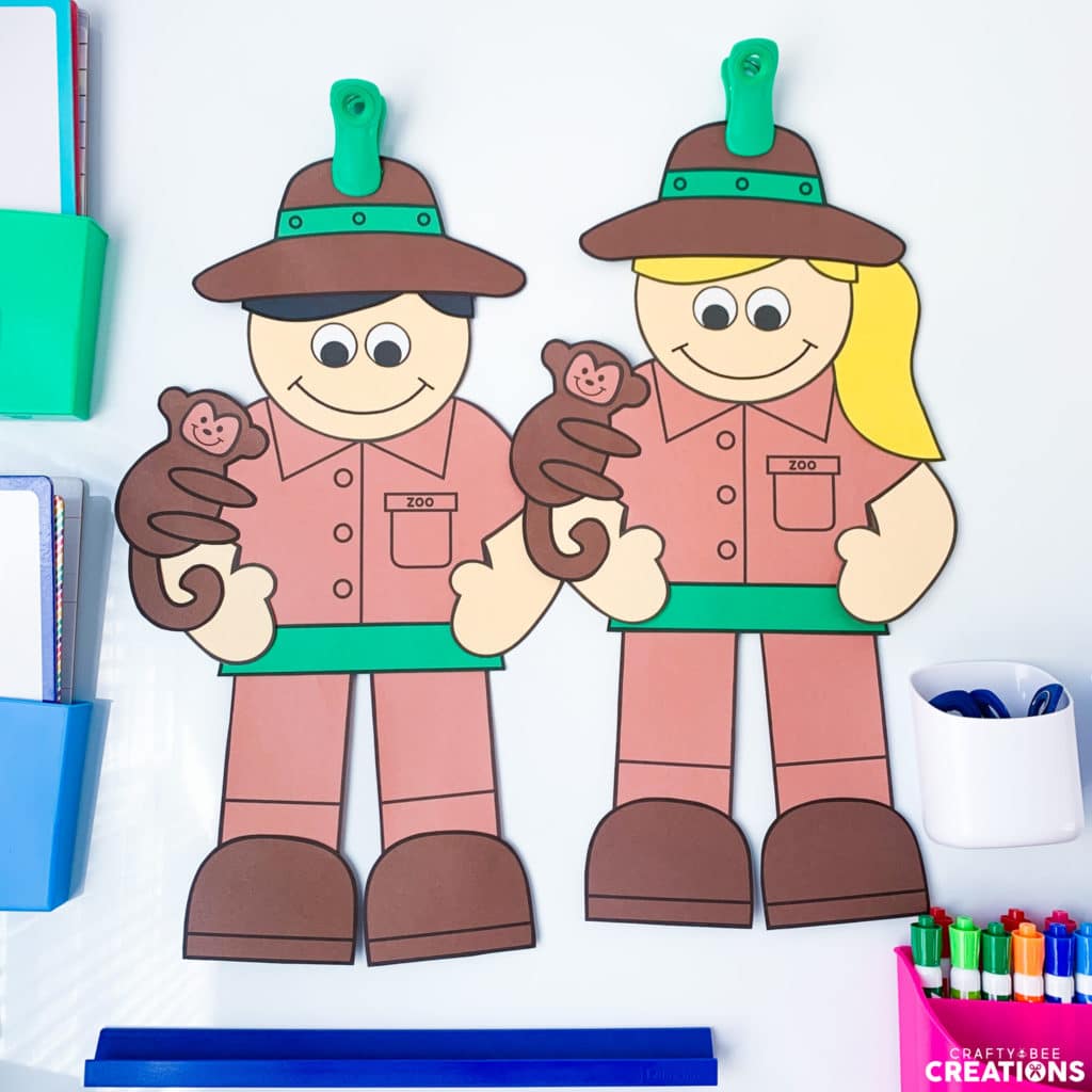 Two zookeeper crafts are hung up by green magnetic clips on a whiteboard. There are dry erase markers in magnetic containers on the board as well. The zookeeper crafts are male and female and are each holding a cute little monkey. The female craft has blonde, yellow hair and the male has black hair.
