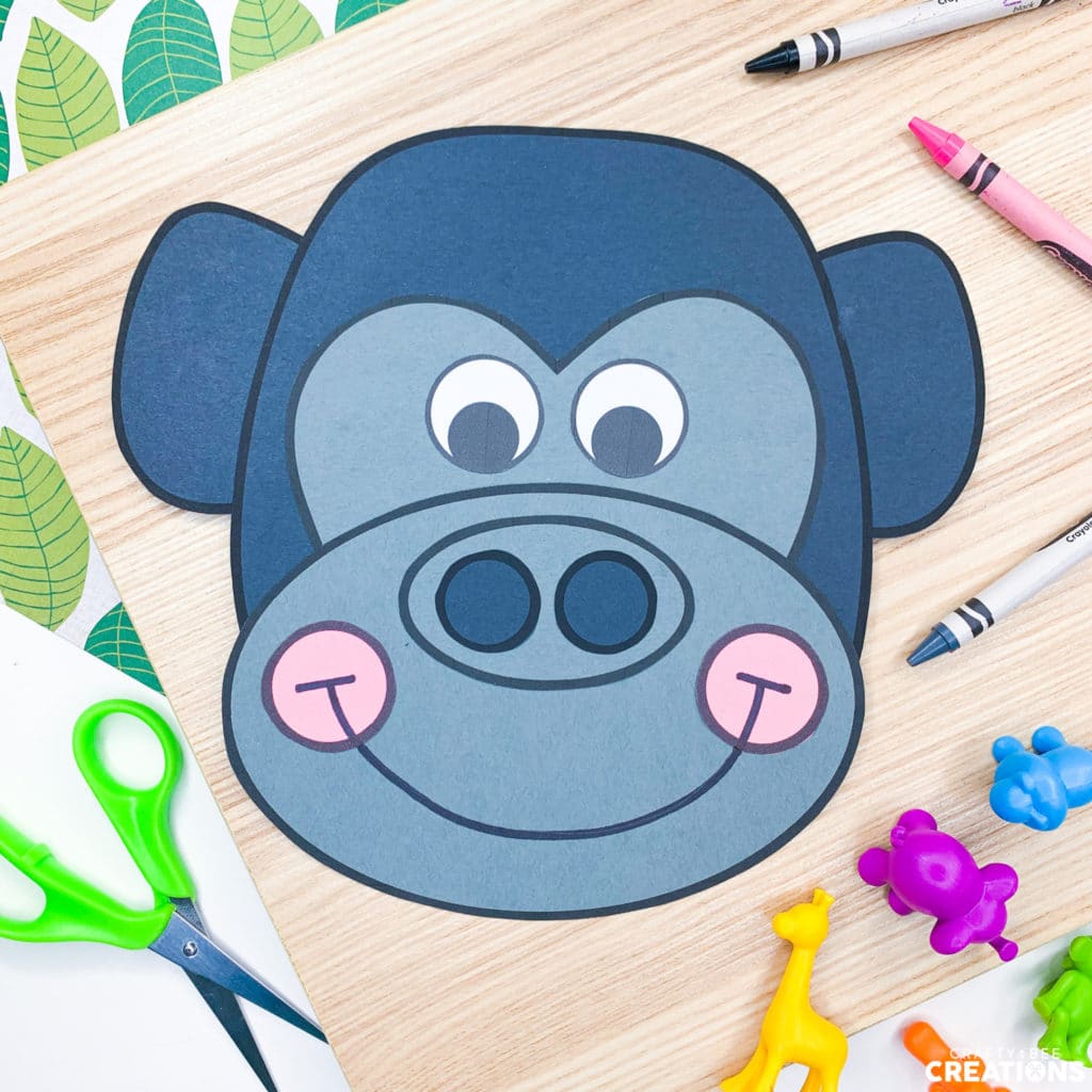 This is a gorilla craft printed on black and gray cardstock paper. There is a gray crayon and some purple and blue and yellow animal figurines lying beside it. There is also a green pair of scissors. The craft is just the gorillas face.