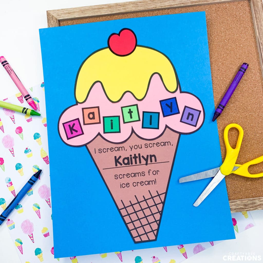 The ice cream cone craft is displayed on blue paper and set on a cork board and sheet of scrapbook paper covered in ice cream treats and cotton candy. There is a yellow pair of scissors as well as a pink, blue, purple and green crayon lying on the page.