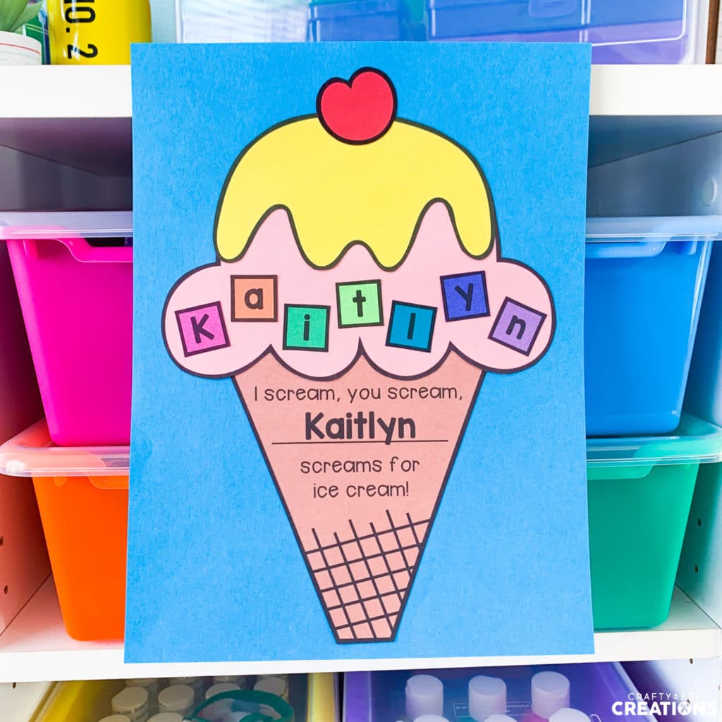 The ice cream cone craft is displayed on blue paper and says the name Kaitlyn. The craft is hung on a manipulative shelf holding colored bins. The cone says, "I scream, you scream, Kaitlyn screams for ice cream!"