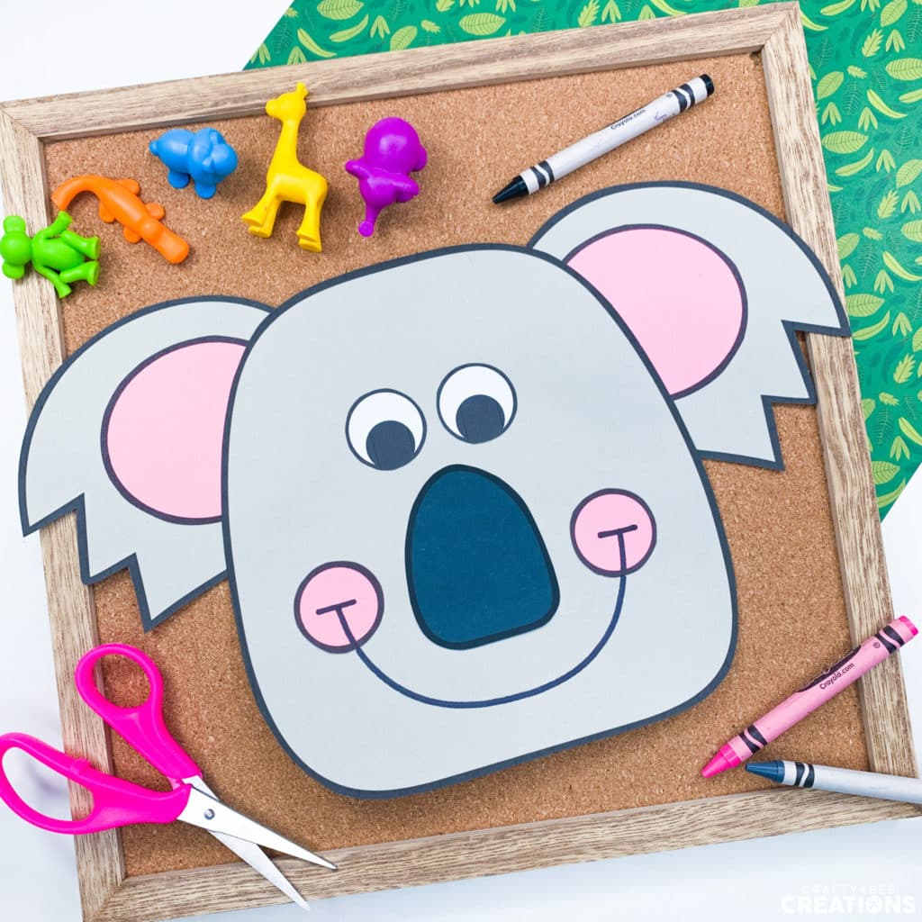 The image shows a gray koala bear, a pair of pink scissors and three crayons. The koala craft is on a small cork board. There are also five colorful animal figurines lying nearby.