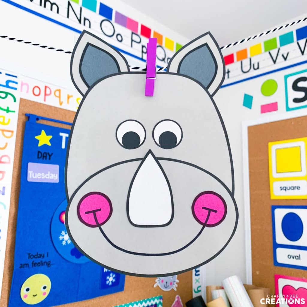 The rhino craft is hanging from a purple clothespin with some classroom decor behind it. The rhino craft has pink cheeks.