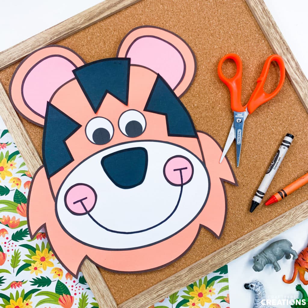 The orange and black tiger craft is lying on a small cork board. There are orange children's scissors and some crayons lying nearby. There is also a hippo, zebra, and tiger figurine by the board.